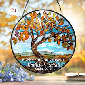 May include: A round stained glass window featuring a tree with orange, yellow, and brown leaves. The text "HAPPY 7TH ANNIVERSARY Mathew & Sarah 08.26.2018" is displayed at the bottom. The window is framed in black and hangs from a chain.
