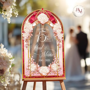 May include: A decorative wedding sign on an easel. The sign has a pink and gold frame with white roses and the text "Celebrating 15 Years of Love" with the names "Paul & Maria" and the years "2010-2025".