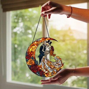 Pode incluir: Apanhador de sol em vitral em forma de lua, apresentando um casal de esqueletos dan&ccedil;ando. A lua &eacute; decorada com rosas vermelhas, amarelas e laranja. Os esqueletos est&atilde;o vestidos com trajes formais, o homem usa uma cartola.