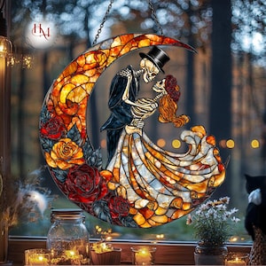 Pode incluir: Vitral em forma de lua crescente com um casal de esqueletos em trajes formais, abra&ccedil;ando-se. A lua &eacute; decorada com rosas vermelhas e laranja. A obra de arte &eacute; suspensa por uma corrente, com um fundo desfocado de &aacute;rvores e luzes.