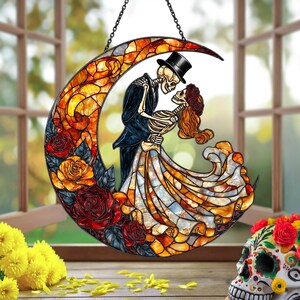 Pode incluir: Suncatcher de vitral com um casal de esqueletos dan&ccedil;ando em frente a uma lua crescente. A lua &eacute; decorada com rosas vermelhas, laranja e amarelas. Os esqueletos est&atilde;o vestidos com trajes formais, o homem usando uma cartola.