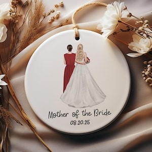 May include: A round, white ceramic ornament featuring a bride and her mother, with the text "Mother of the Bride" and the date "08.20.25". It has a string for hanging. The background is a soft beige.