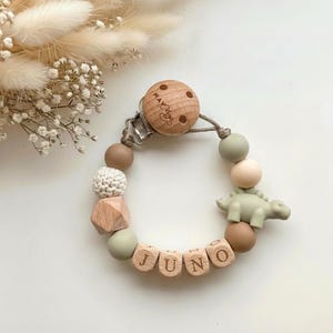 Puede incluir: Un sujetachupetes con un disco de madera grabado, un dinosaurio de silicona verde salvia y cuentas en tonos beige, marrón y verde salvia. Cuatro cuentas de madera en forma de cubo deletrean el nombre JUNO con letras negras grabadas.