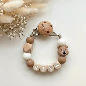 May include: A pacifier clip featuring a round wooden piece engraved with the text May May, connected to a string of beads in white, tan, and brown. The beads include a geometric wooden shape, a bear-shaped bead, and letter beads spelling ZIEN.