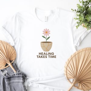 Puede incluir: Camiseta blanca con un estampado floral. El diseño presenta una flor rosa en una maceta agrietada con el texto "HEALING TAKES TIME" en marrón. La camiseta se muestra con abanicos decorativos y una planta.