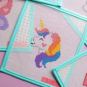 May include: Needlepoint canvases featuring a colorful unicorn design. The unicorn has a rainbow mane and tail, with a yellow horn. The canvases are square, with a white mesh background and a teal border. The brand name "SALT + SAND" is visible.