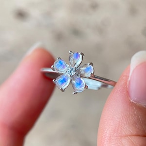Puede incluir: Un anillo de plata con un diseño floral. La flor está compuesta por seis piedras preciosas azul claro iridiscentes en forma de pera, que rodean una pequeña piedra preciosa transparente en el centro. El anillo se sostiene entre dos dedos.