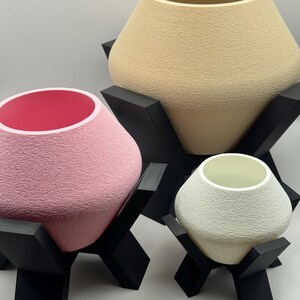 May include: Three tapered, textured pots in beige, pink, and white, each resting on a black, angular, four-legged stand. The pink pot features a bright pink interior, while the beige and white pots have interiors matching their exterior tones.