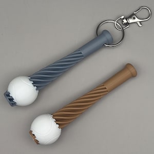 May include: Two baseball bat-shaped keychains. One is gray with a white baseball, the other is brown with a white baseball. Both have a twisted handle design and a metal clasp for attaching to a bag or keys.