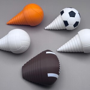 May include: Five sports-themed bottle stoppers. The stoppers are shaped like a basketball, soccer ball, volleyball, baseball, and football. The basketball stopper is orange, the soccer ball is black and white, the volleyball and baseball are white, and the football is brown.