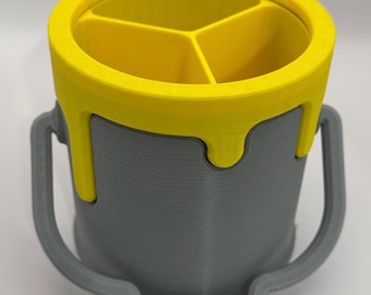 May include: A grey cylindrical desk organiser shaped like a paint bucket with a yellow rim featuring a dripping paint design. The interior is divided into three sections by yellow partitions, and the exterior includes a grey handle.