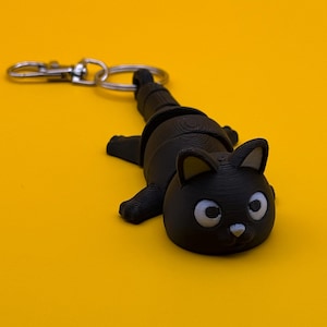 May include: A black cat-shaped keychain with a silver clasp. The cat has large, round eyes and a small nose. The keychain is made of a solid, matte black material and is lying on a yellow surface.