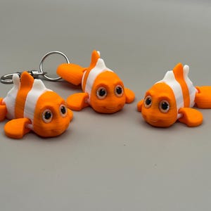 May include: Three orange and white clownfish keychains with a silver keyring. The fish have large, expressive eyes and are lying on a gray surface. The keychains are a fun, whimsical accessory.