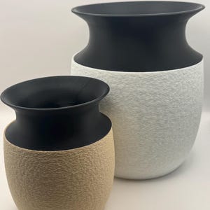 May include: Two decorative vases with a textured surface. The larger vase is white with a black top, and the smaller vase is tan with a black top. The vases have a modern, minimalist design.