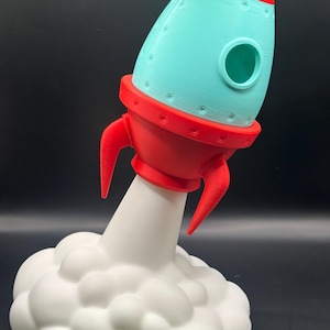May include: A 3D-printed rocket ship toy in red and teal, with a white cloud base. The rocket has a cone-shaped top, a circular window, and red fins. The cloud base is a fluffy, rounded shape.