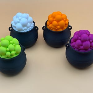 May include: Four black cauldron-shaped containers, each filled with a different color of bubbly, spherical foam. Colors include white, orange, green, and purple. The cauldrons have small handles on either side and sit on a light tan surface.