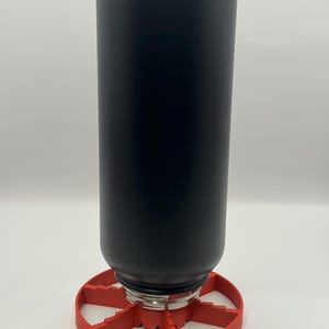 May include: A black insulated water bottle with a silver base is set in a red, circular holder. The holder has a starburst design. The bottle is cylindrical and appears to be made of metal.
