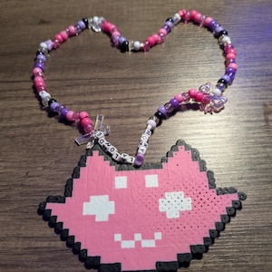 May include: A handmade necklace featuring a pink pixelated cat pendant and a beaded chain. The chain is made of pink, purple, and clear beads, with a butterfly charm. The pendant has white pixelated eyes and mouth. The necklace has the text "LA LONDE" and "ROXY".