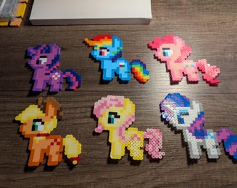 May include: Six colorful pixel art pony figures in various colors, including purple, blue, pink, yellow, orange, and white. Each pony has a unique design and color scheme, with details like manes, tails, and cutie marks. They are arranged on a wooden surface.