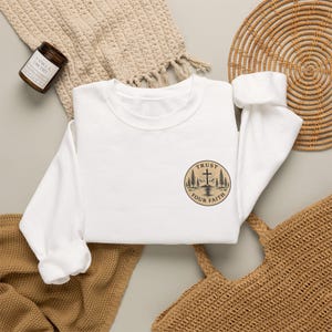 May include: A white long-sleeve sweatshirt featuring a circular graphic on the chest. The design depicts a cross standing on a hill surrounded by pine trees and a sunburst, with the text "TRUST YOUR FAITH" printed along the circular border.