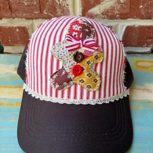 May include: A baseball cap with a dark brown brim and a white and red striped front panel. A fabric gingerbread person applique with floral and plaid patterns is centered on the front, accented by a small red and white striped bow and two buttons.