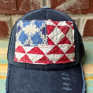 May include: A dark navy blue baseball cap with a distressed, frayed brim and a rectangular patch on the front. The patch features a blue section with a white floral pattern and a red and white quilted diamond pattern resembling a flag design.