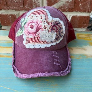 May include: A distressed purple trucker hat with a heart-shaped patch. The patch features a floral design, lace trim, and the word "love." The hat has a mesh back and a frayed brim.
