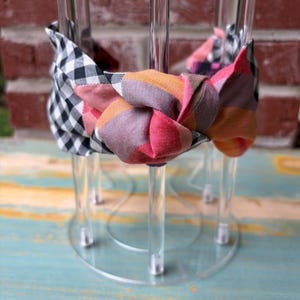 May include: A fabric headband with a knotted bow. The bow features a mix of patterns and colors, including black and white gingham, pink, orange, and gray plaid. The headband is displayed on a clear acrylic stand.