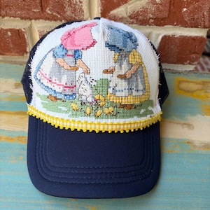 May include: A navy blue baseball cap featuring a white front panel with a cross-stitch embroidery design of two figures in bonnets feeding chickens and chicks. The cap is accented with a yellow and white checkered trim along the base.