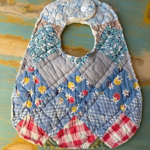 May include: A handmade baby bib with a patchwork design. The bib features various blue fabrics with floral, striped, and checkered patterns, along with red and white gingham. It has a round neck opening and a white snap closure.