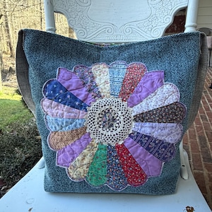 May include: A large, handmade tote bag with a denim-like texture. The bag features a colorful, quilted flower design in shades of purple, blue, green, and red, with a cream-colored crocheted center. The bag has brown straps.