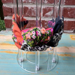 May include: A clear acrylic headband display stand holding three fabric headbands. The headbands feature floral, paisley, and geometric patterns in orange, black, pink, and white. The stand is on a light blue surface with a brick background.