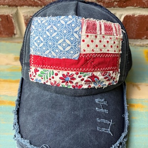 May include: A dark blue distressed baseball cap featuring a rectangular patch on the front panel. The patch is a patchwork design resembling a flag, composed of blue geometric patterns, red and white stripes, and a floral print fabric.