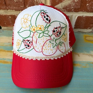 May include: A red and white baseball cap featuring an embroidered design on the front panel. The embroidery depicts strawberries, yellow flowers, and ladybugs in red, green, and yellow thread. A delicate cream-colored lace trim borders the bottom.