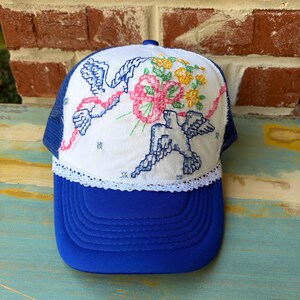 May include: A royal blue trucker hat featuring a white front panel with embroidered blue birds, a pink ribbon, and yellow flowers. The hat includes a white lace trim along the base of the front panel and a blue mesh back with a solid blue brim.