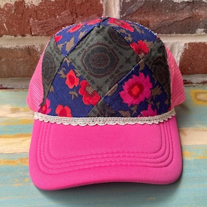 May include: A pink trucker hat with a floral patchwork design on the crown. The patchwork features blue fabric with red flowers and a green and brown pattern. The hat has a pink mesh back and a white lace trim.