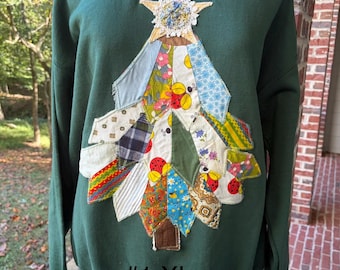May include: A dark green long-sleeve sweatshirt featuring a large, central Christmas tree applique made from various colorful fabric scraps with floral, plaid, and ladybug patterns. A textured star sits at the top. The text #1 XL is printed below.
