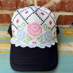 May include: A black baseball cap featuring a white front panel with a cross-stitch floral pattern in pink, blue, and green. The white panel is accented with a black diamond grid design and a white crocheted lace trim along the bottom edge.