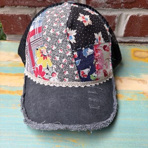 May include: A patchwork baseball cap with a black mesh back and a denim brim. The crown features various floral and patterned fabrics in red, white, black, and blue. The brim is a faded gray denim with a frayed edge.