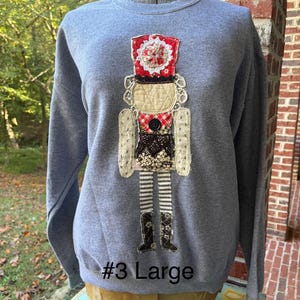 May include: A heather gray crewneck sweatshirt featuring a central applique of a nutcracker figure. The nutcracker is constructed from various fabric patches, including red, white, and black lace, gingham, and striped patterns. The text #3 Large is visible.