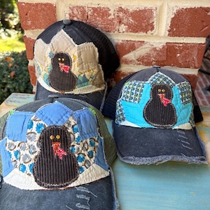 May include: Three baseball caps featuring a dark gray distressed brim and a black mesh back. Each cap displays a central applique of a dark brown corduroy turkey with a red patterned wattle, surrounded by various colorful fabric quilt patches.