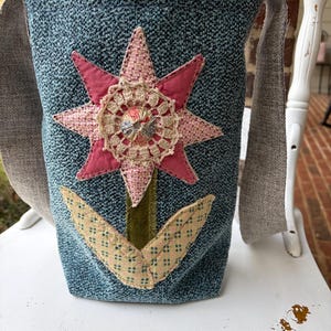 May include: A teal fabric tote bag with a large, appliquéd flower design. The flower features a pink star-shaped center, a crocheted center, and green and yellow leaves. The bag has gray fabric straps.