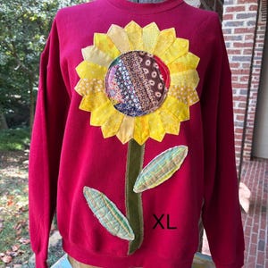 May include: A red long-sleeve sweatshirt featuring a large, multi-textured sunflower appliqué on the front. The flower has yellow fabric petals and a dark, patterned center. A green stem and two green leaves are attached. The text XL is visible.