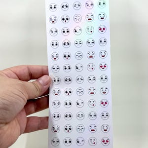 May include: A sheet of stickers with various emoji faces, held by a hand. The stickers are white with black outlines and feature different expressions. The text "Emo-tate" and "Emojis to annotate your books" are at the top.
