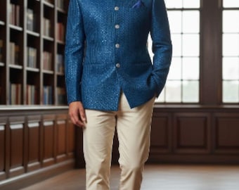 Puede incluir: Un hombre con un atuendo formal, con una chaqueta azul texturizada con cuello mandarín, pantalones claros y zapatos de cuero marrón. La chaqueta tiene botones plateados y un pañuelo de bolsillo. El fondo es una biblioteca con estanterías y ventanas grandes.