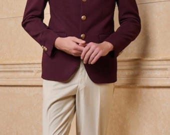 May include: A burgundy blazer with gold buttons and a decorative pin, paired with beige pants. The outfit is from King Men's Tailor. The blazer has a mandarin collar. The pants are a neutral color.