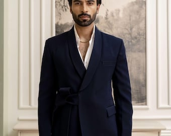 May include: A navy blue suit with a shawl collar and a matching belt tied at the waist. The suit is paired with a white dress shirt and a gold chain. The background features a painting and a white cabinet.