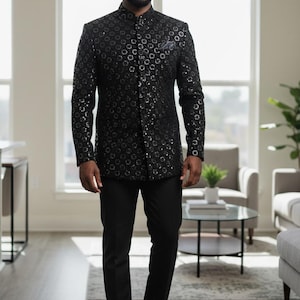 Op de afbeelding: Een zwart jasje met patroon, een opstaande kraag en een bijpassende zwarte broek. De jas heeft een zich herhalend cirkelvormig ontwerp. De outfit wordt gecompleteerd met zwarte geklede schoenen. De persoon staat in een goed verlichte kamer.