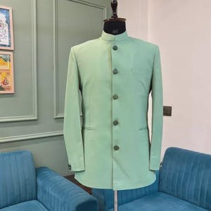 May include: A light green suit jacket with a mandarin collar and matching buttons. The jacket is displayed on a mannequin with a size 40 label. The jacket has a pocket on the left side. The background includes teal blue couches and a light green wall.