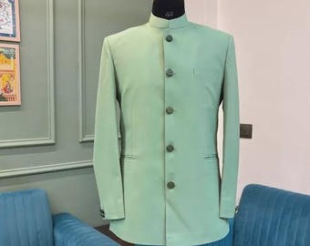 Puede incluir: Chaqueta de traje verde claro con cuello mandarín y botones a juego. La chaqueta se exhibe en un maniquí con una etiqueta de talla 40. La chaqueta tiene un bolsillo en el lado izquierdo. El fondo incluye sofás azul verdoso y una pared verde claro.